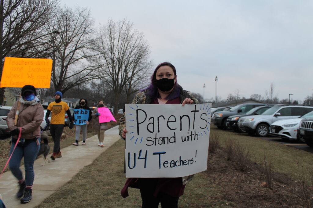 A woman holds a sign that reads "Parents stand with U4 Teachers." Next to her, other parents and teachers march with signs.