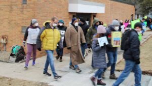 A man with a tan pea coat walks in the middle of a crowd of blurrier marchers, in front of a brick building. 