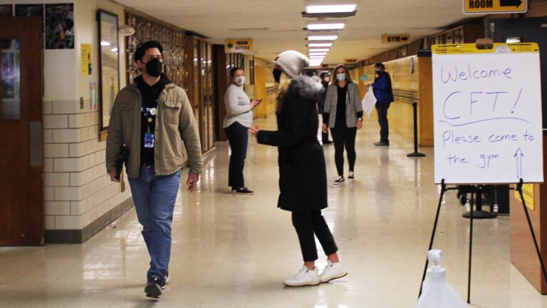 A white man in a tan coat walks in a yellow school hallway.