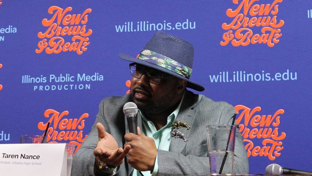A man in a blue hat and a blazer speaks into a microphone. 
