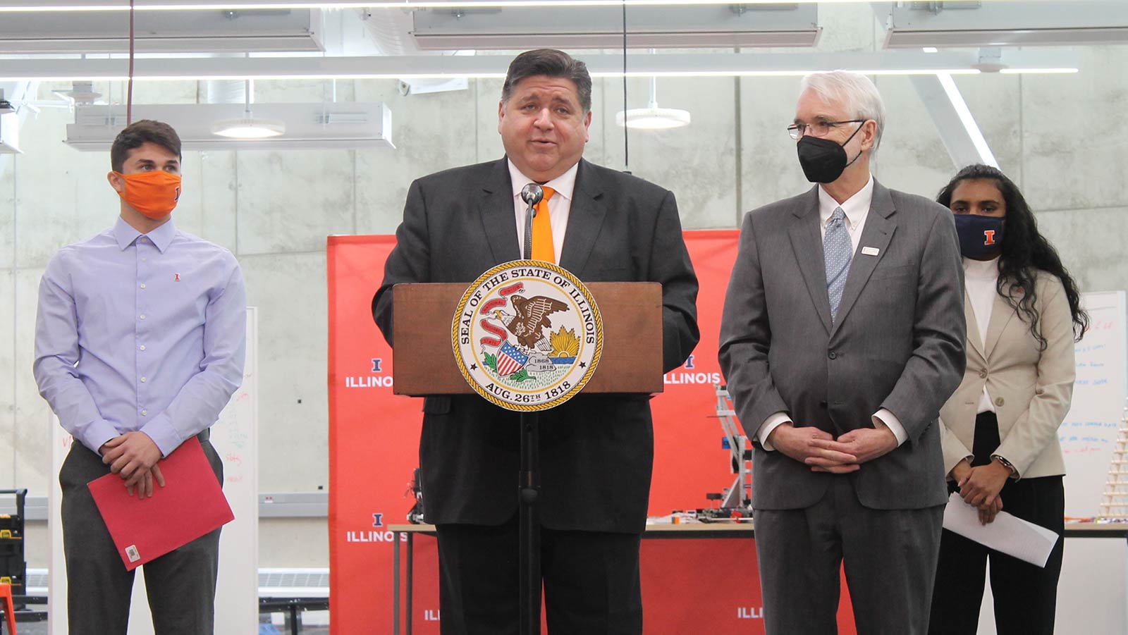 The governor stands at a podium, flanked by two students and an elderly man.