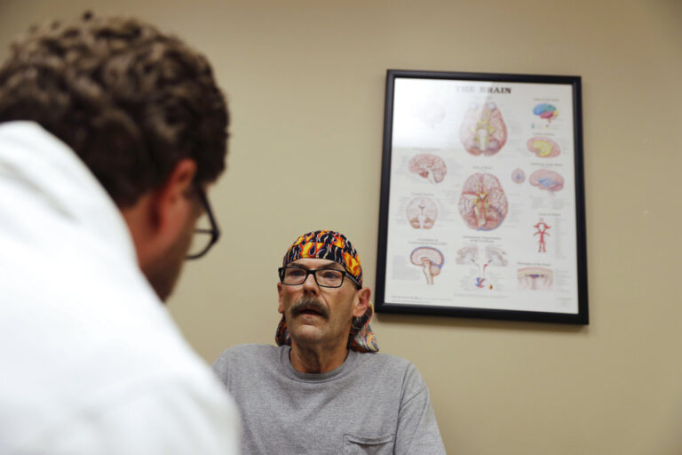 A man sits next to a doctor's office brain chart. In the foreground, a man wears a white coat.