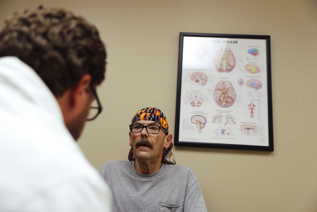 A man sits next to a doctor's office brain chart. In the foreground, a man wears a white coat.
