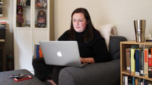A woman sits cross-legged in a sofa chair. She types on a laptop. A bookshelf is on her left.