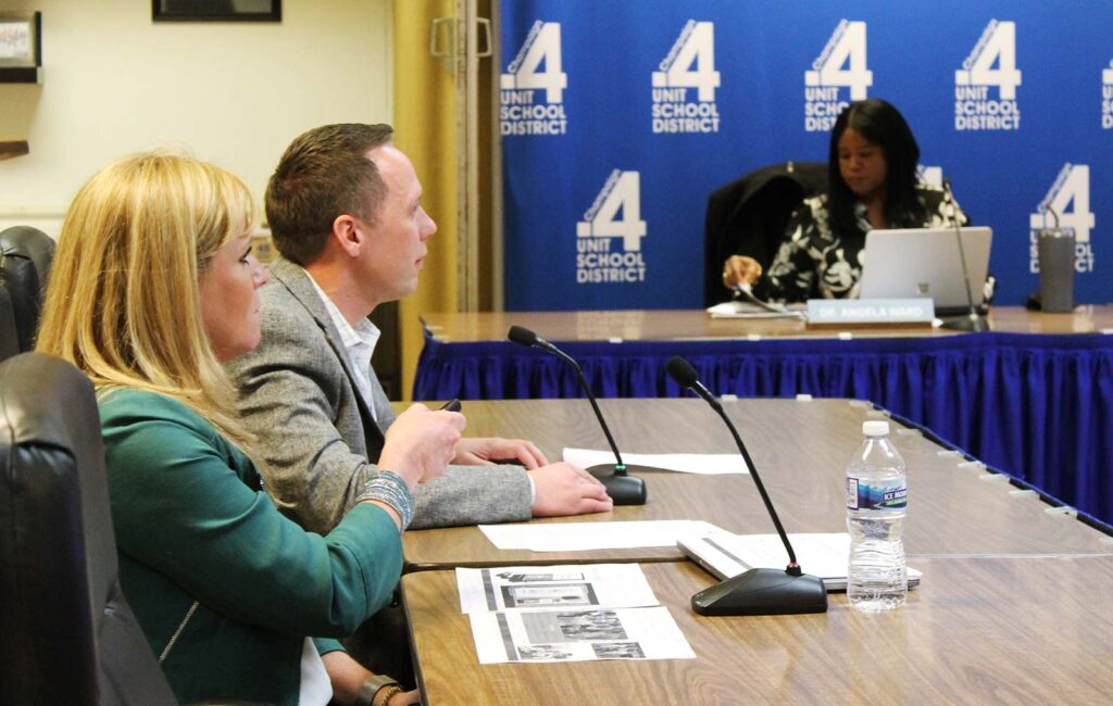 A woman holds a clicker. She's sitting next to a man at a Board of Education meeting.