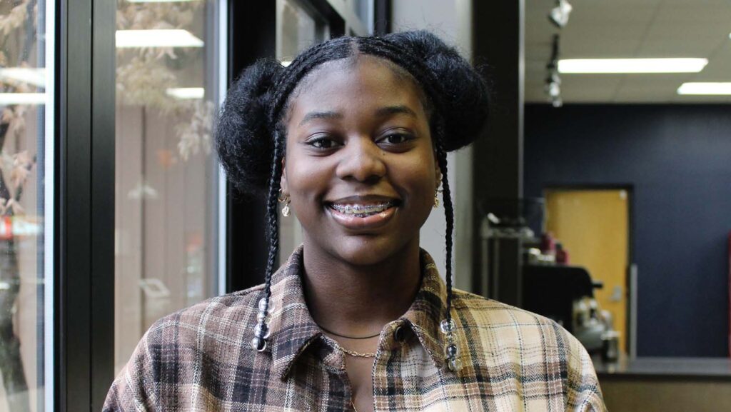 A young woman with Afro puffs and a plaid shirt smiles at the camera.