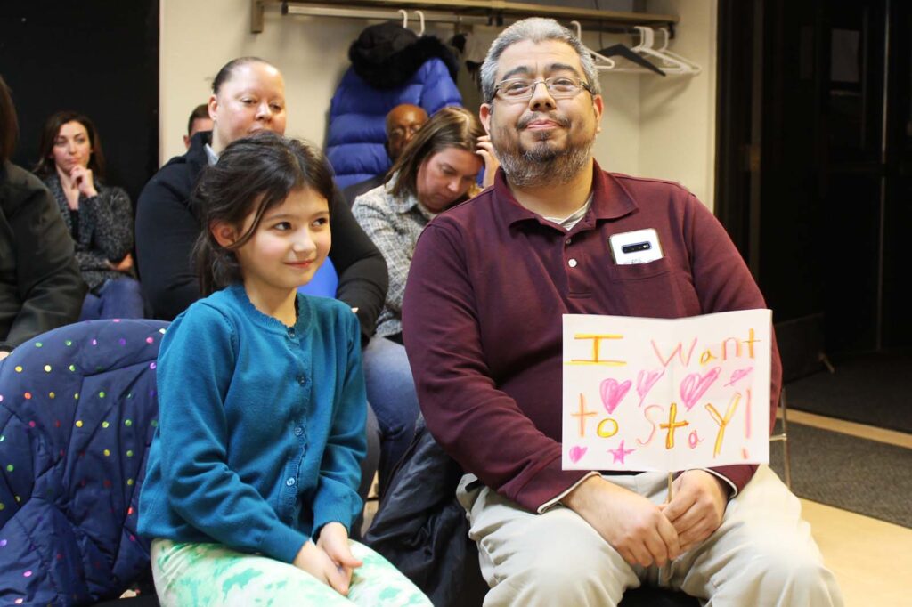 A little girl sits next to her father, who holds a sign that reads "I want to stay," and is decorated with hearts.